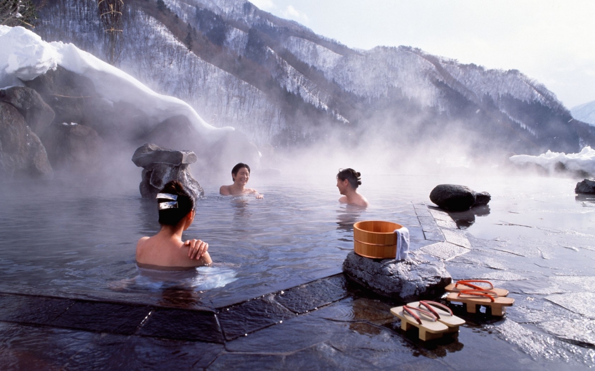 Three people relax in a hot spring, surrounded by snow-covered mountains in the background