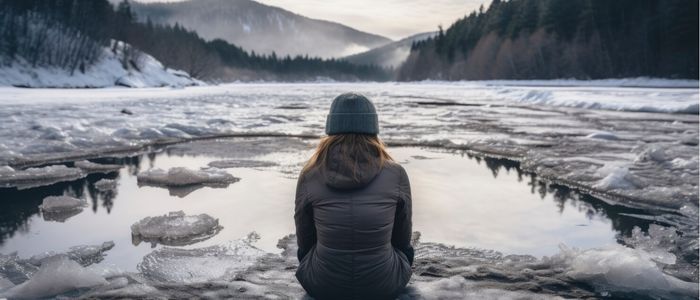 A person in a winter coat and beanie sits on icy ground, facing a frozen lake surrounded by snow-covered hills and pine trees under a cloudy sky.