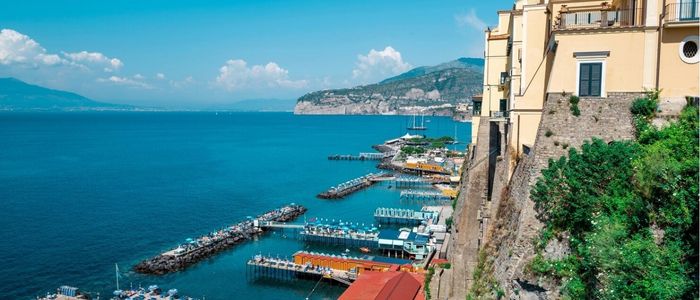 A person in a winter coat and beanie sits on icy ground, facing a frozen lake surrounded by snow-covered hills and pine trees under a cloudy sky.