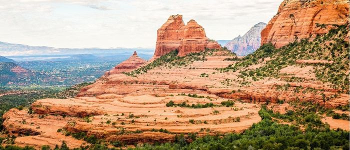 Red rock formations in Sedona, Arizona, under a cloudy sky. Layered rock and sparse greenery create a rugged, tranquil landscape with distant mountains.